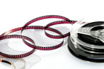 Valokuva filmikelasta, jossa filminauhaa purkautunut. Nauha on punertavaa ja kelan kohdalta mustaa. Kuvan tausta on valkoinen. Filmikela muovia.